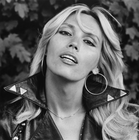 Portrait of Amanda Lear, mouth open, wearing hoops and leather jacket.