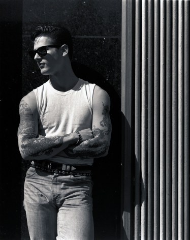 Man in a white tank top, jeans and sunglasses leaning against a wall with arms crossed.