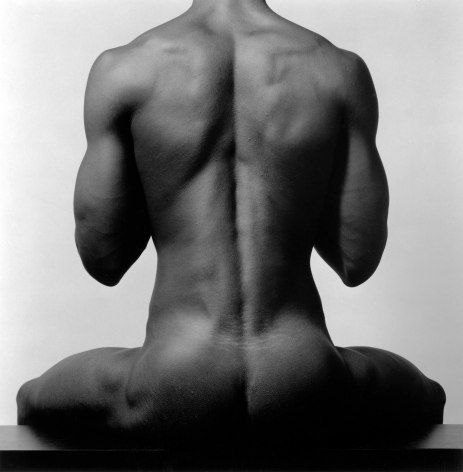 Nude black man from behind sitting upright.