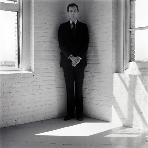 Robert Wilson wearing a suit standing in the corner of a white room.