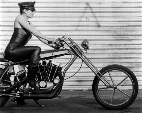 Lisa Lyon in leather outfit and hat, on motorbike.