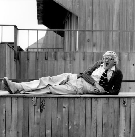 Portrait of David Hockney reclining on wooden bench and yawning.