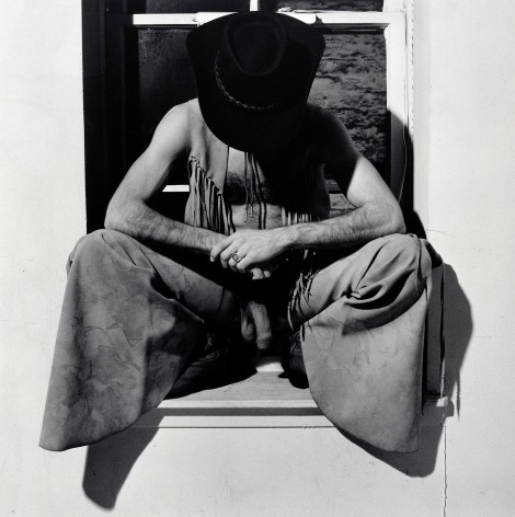 Man crouching in window, wearing cowboy hat, pants, and and vest looking down with penis visible.