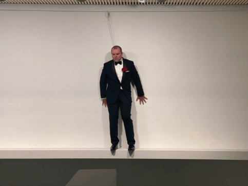 Art performance of a man in a tuxedo standing on a short ledge in a gallery