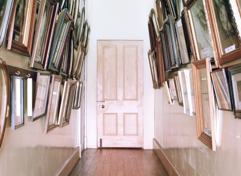 Narrow hallway with wood floors and door at the end, hung with many framed pictures on both sides of the passage.