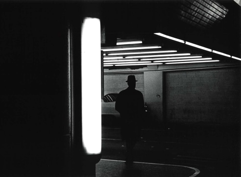 Black and white photo showing the silhouette of man in a hat walking through a parking garage.