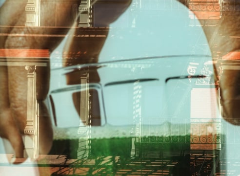 Color photo of a close up of an hand holding a glass of green juice superimposed on a reflection of a building.