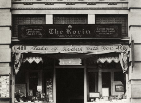 Black and white photo of the The Koran, a storefront Kodak Store based in LA in the 1920s—framed pictures are in the shop windows and there is an awning that reads "Take a Kodak With You"