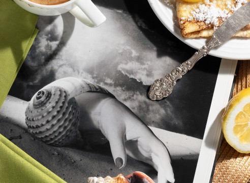 Color photograph of a breakfast table with coffee, a crepe, and a book opened to a surrealistic black and white photo of a hand emerging from a shell.