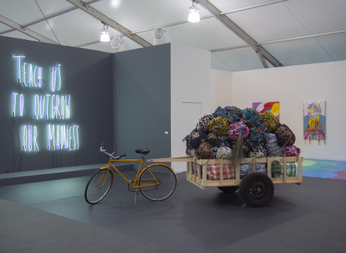 Art fair booth featuring neon text, bicycle sculpture, and paintings