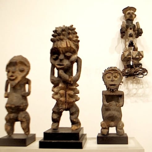 Four wood sculptures of humanoid, geometric shapes face us directly. Three of them are standing on a white block mount, and another is mounted on the white wall to the left. 