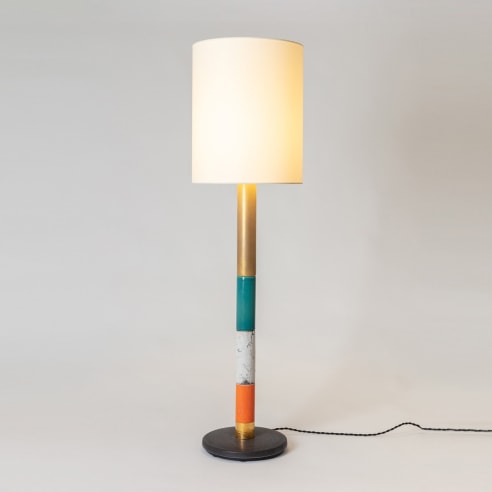 Floor lamp in various colors, white fabric shade. Colors from bottom to top, black base, gold section, orange section, white, green raku, gold. Light turned on. 