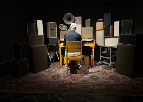 Person sitting at an organ in front of over a dozen mixed speakers 