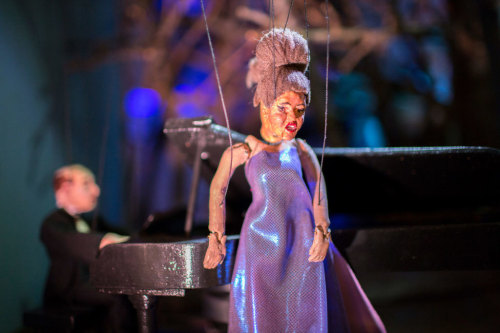 Marionette singer standing in front of a piano