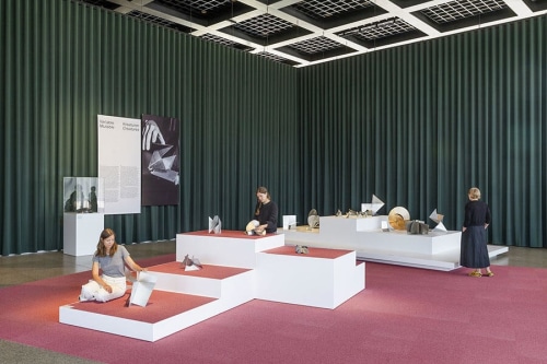 Museum installation of sculptures on top of white plinths with pink carpeting and green curtains