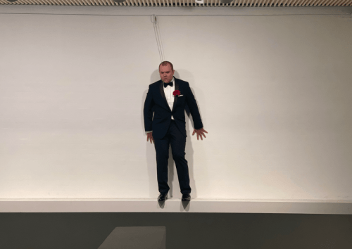 Art performance of a man in a tuxedo standing on a short ledge in a gallery