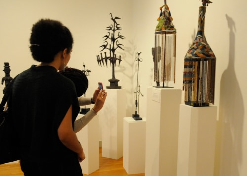 Two adults with their backs to the viewer, taking photos and gazing at Nigerian sculptures, which sit on top of white pedestals, placed in the corner of two white walls, to the right of the image.