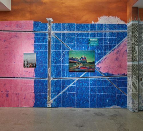 this is an image of the exhibition SUPERUNKNOWN HELD AT Nahmad Contemporary. It features as a wall paper a gigantic photo by Urs Fischer which represents loosely a construction site,  scaffolding and a fence, the colors are rich, pink on the left and dark blue on the right.  Two small paintings are featured in the center on bey Yves Tanguy and the other by Max Ernst.