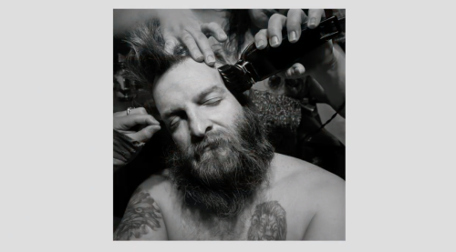 A bearded man has his long hair shaved in this black and white photo