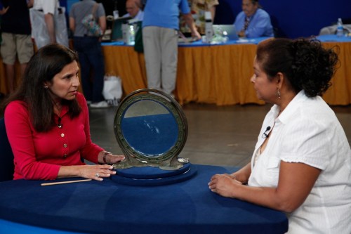 At an Antiques Roadshow appraisal event, Arlie Sulka and a guest sit at a table discussing a round dressing mirror by Tiffany Studios with peacock feather motif.