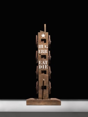 The sculpture The American Dream, made of a beam with a haunched tenon, with the words Hug, Err, Eat, and Die painted in white, and a white star painted on top. Eight wheels have been attached to the sculpture, four on the right and four on the left.