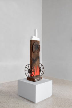 Indiana's sculpture soul, consisting of a salvaged wooden beam atop a wood base with the title Soul painted in white stenciled letters. Red flames are painted at the bottom of the beam and a metal wheel is attached to each side. At the top of the work is a circular metal fixture, and the haunched tenon is painted white. 
