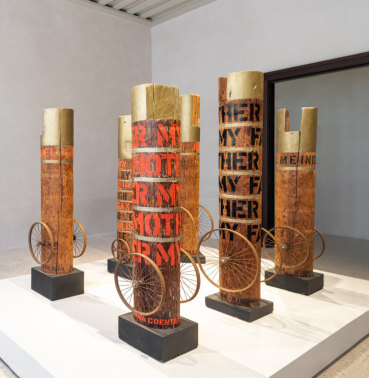 Installation view of six of Indiana's columns, all have two wheels attached to the bottoms sides, text, and gold tops