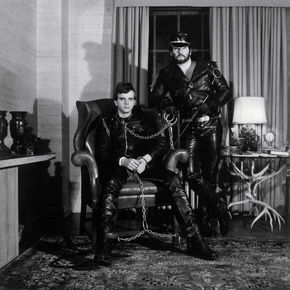 Lyle Heeter and Brian Ridley in full leather and a harness, seated in their living room.