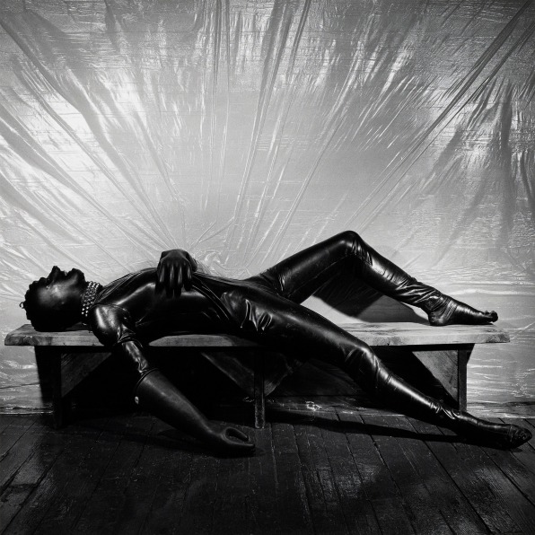 man in full rubber suit reclining on a bench in front of a plastic sheet.