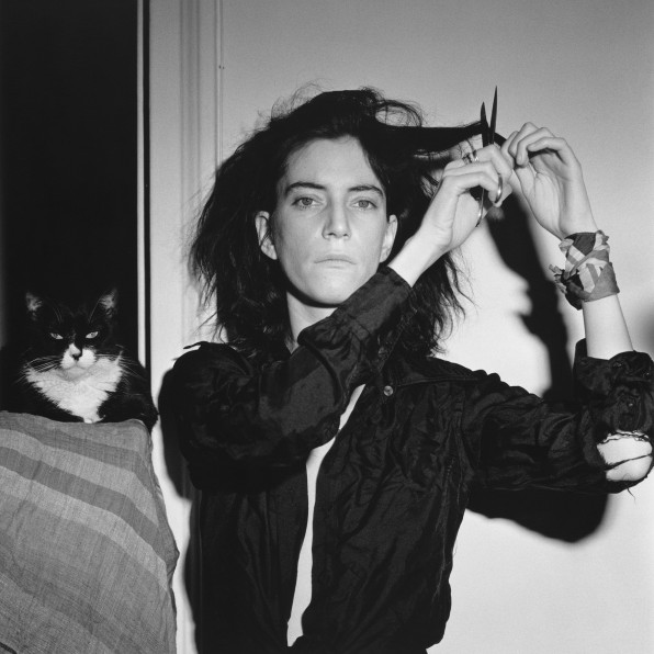 Patti Smith facing camera, cutting her hair, with cat in background.