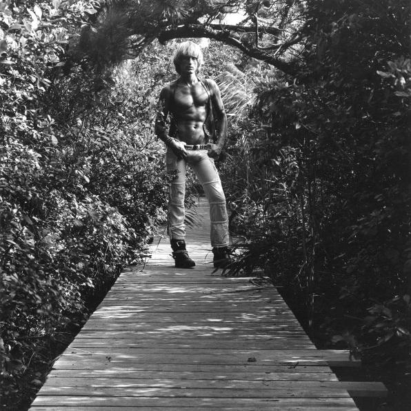 Peter Berlin, shirtless, with hands on hips, standing on boardwalk surrounded by foliage.