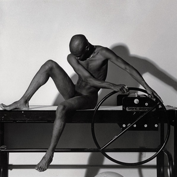 Nude black man sitting with hands on the wheel of a slab roller and looking downward. 