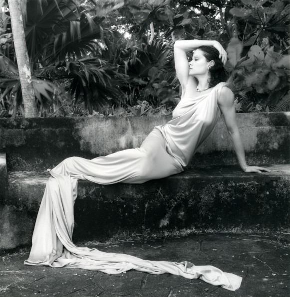 Lisa Lyon in silk dress reclining on a stone bench in a tropical landscape.