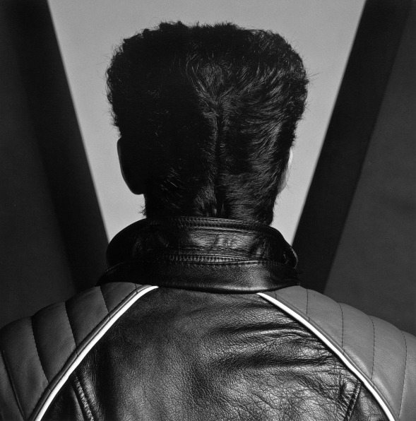 A man photographed from behind standing in front of a geometric backdrop. He wears a leather jacket and the hair at the back of his head is parted.