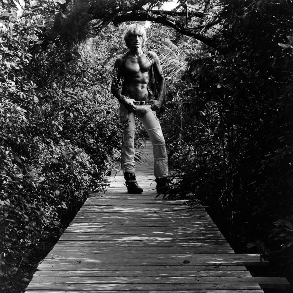 Peter Berlin, shirtless, with hands on hips, standing on boardwalk surrounded by foliage.