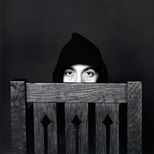 Portrait of Lucio Amelio in a hood hiding behind a wooden chair.