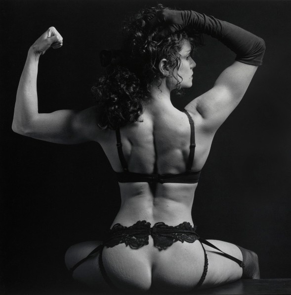 Woman wearing lace lingerie photographed with her back to the camera. She wears an opera glove on her right arm and her left arm is flexed.