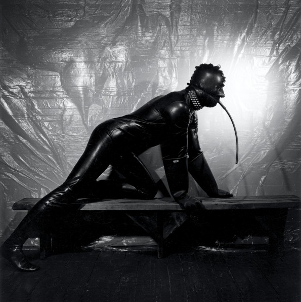 Man in full rubber suit with a hose in his mouth kneeling  on a bench in front of a plastic sheet.