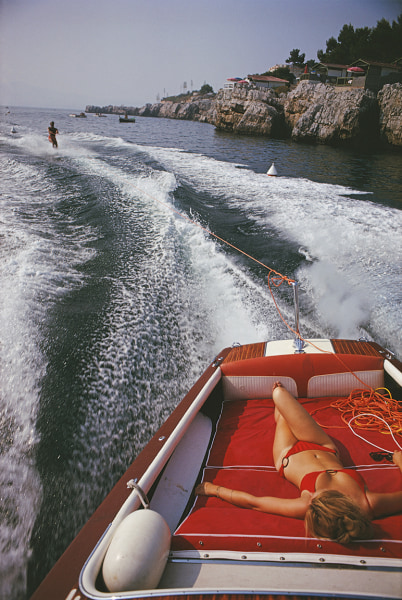 Slim Aarons, Leisure in Antibes: A woman sunbathes on a motorboat as it tows a waterskier in the bay off the Eden-Roc, 1969