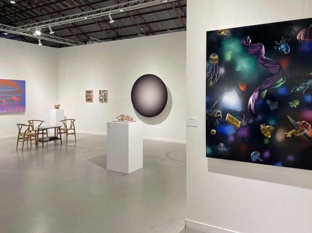 An art fair booth featuring paintings of varying sizes, a sculpture on a pedestal, a table and chairs, and a large, cirucular art piece.