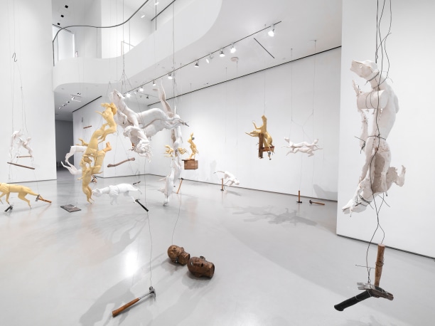 an artist studio with sculptures of animals hanging from the ceiling, an assortment of boxes, art materials, and chairs and desk off to the right with a video projection on a portion of the back wall