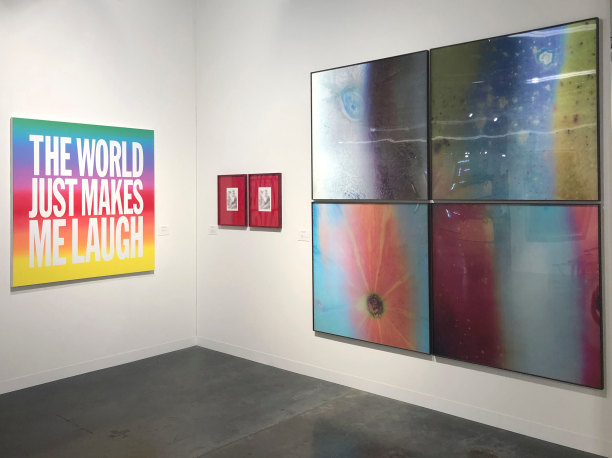 Three artworks hang in a gallery. A rainbow painting by John Giorno reads "THE WORLD JUST MAKES ME LAUGH". There are holographic, colorful abstract photographs by Jitish Kallat and a red diptych by Andrew Sendor.