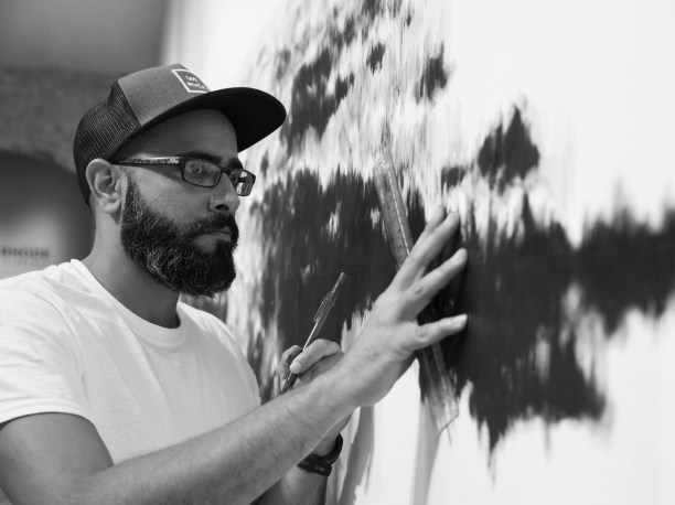 portrait of Gamaliel Rodríguez wearing glasses and a trucker hat and holding a ballpoint pen and ruler up to a work of art