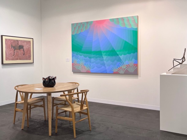 A tradeshow booth with white walls, on the left is a framed artwork of a black horse with a clay red background, on middle wall a blue and green and pink painting of a sun emerging from clouds, and on the right a pedestal with a sculpture of a maquette. In the center, a circular wooden table and three wooden chairs with a black ceramic sculpture of a purse on the tabletop 