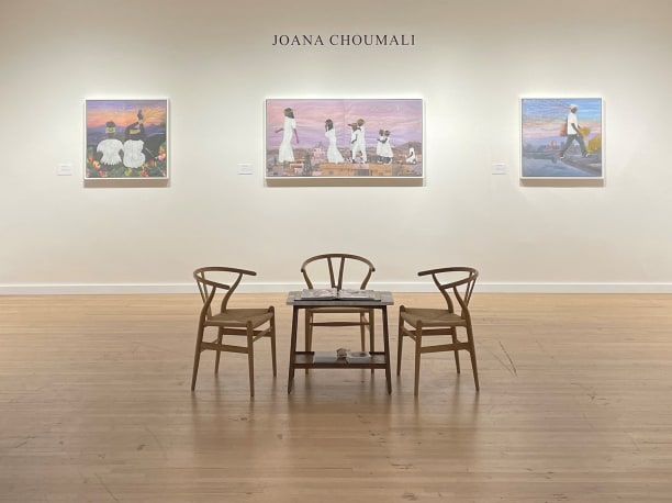 installation view of an art fair booth with a table and three chairs in the foreground and three artworks on the wall behind