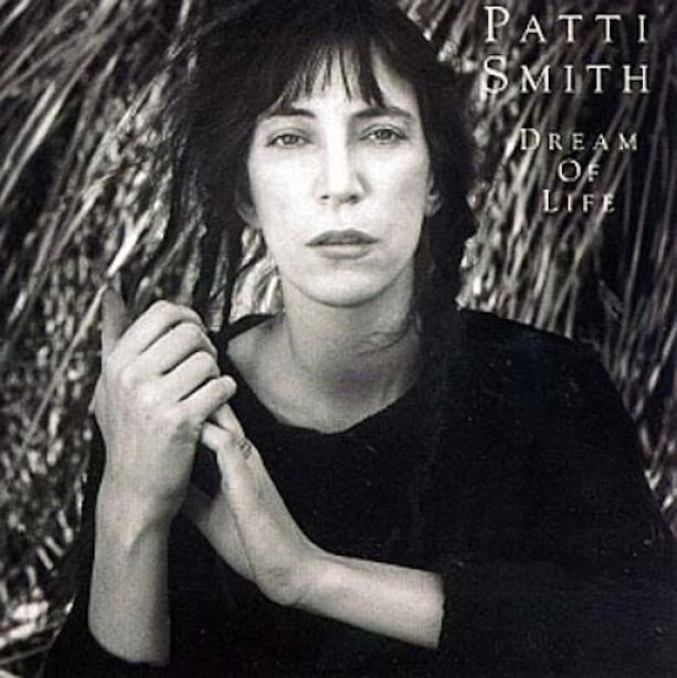 Patti Smith Dream of life album cover, portrait of Patti with hands clasped, looking at camera, hair in braids.