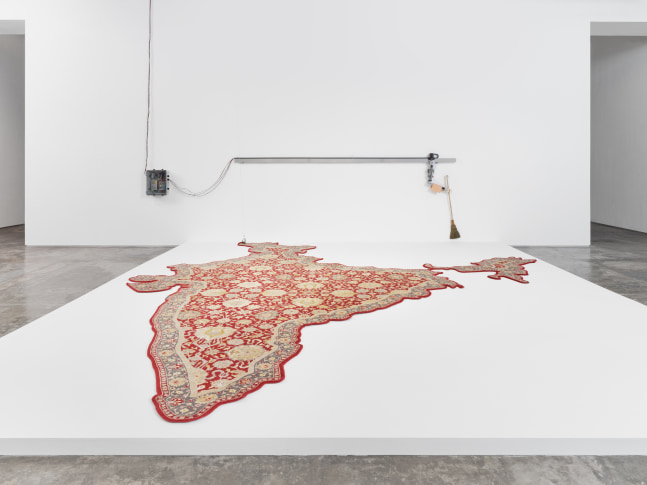 A large woven rug in the shape of post-partition India with the corner attached to a pulley that raises the corner of the rug when a robotic wall mounted arm carrying a broom approaches and sweeps beneath it before gliding back along it's track.