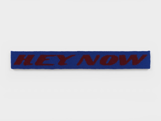 A knit work rendered in blue and burgundy yarn that reads &quot;Hey Now&quot; in a long thin rectangle sewn into linen.