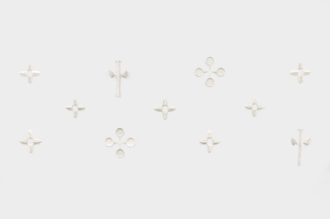 A multi piece wall mounted sculpture with ceramic replicas of irons, cast iron pans and rakes hung in groups of three and four along the wall making a star like pattern.