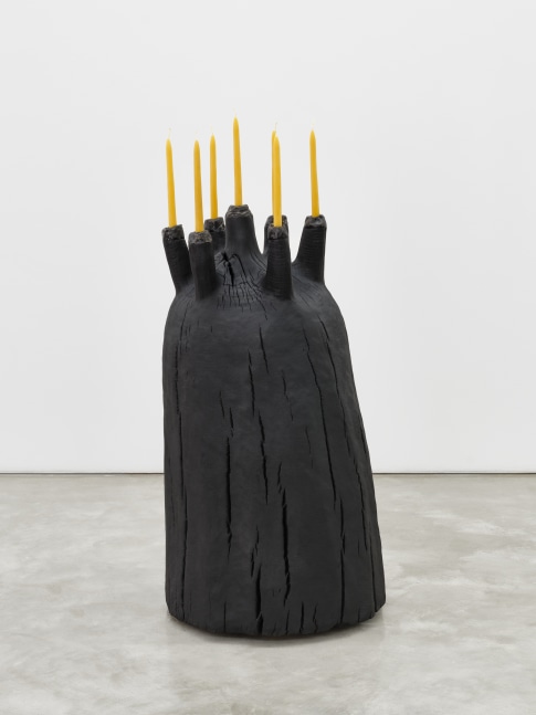 A black mound shaped wooden sculpture with seven long taper candles protruding from the top.